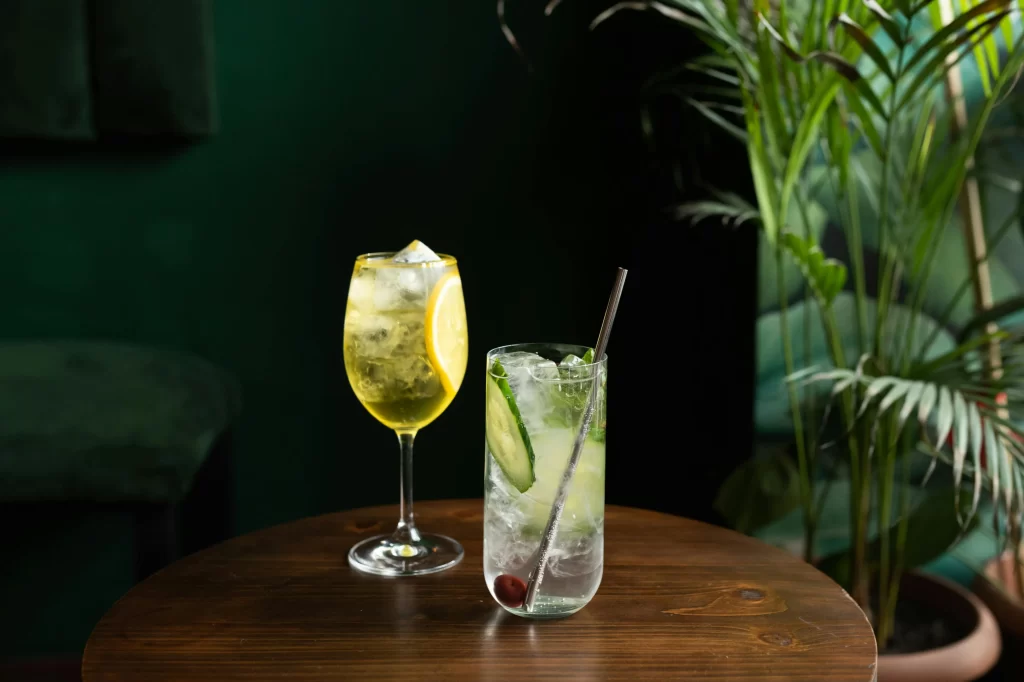 Two cocktails sit on a dark wooden table by potted palms.