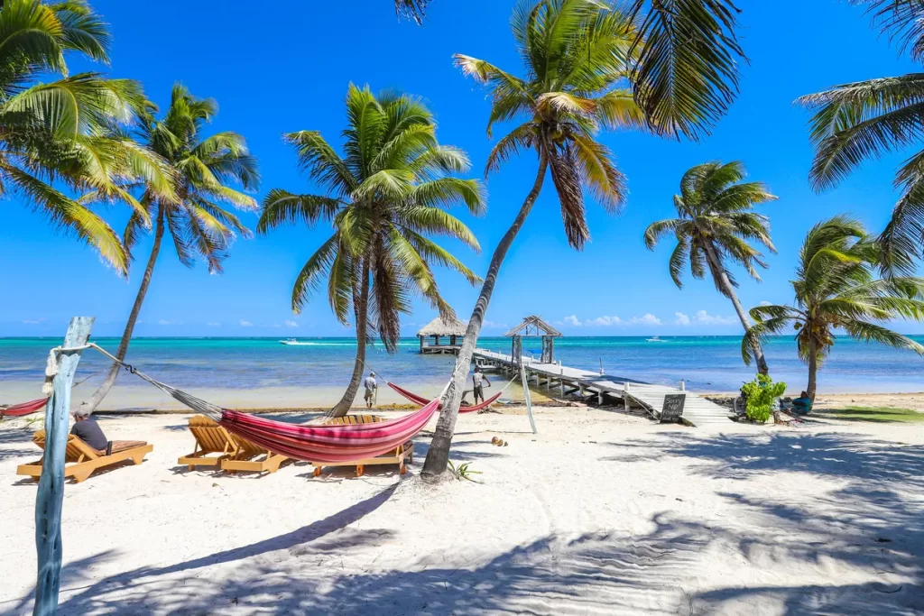 A beach scene in Ambergris Caye, Belize, with wooden lounge chairs, a striped hammock, and wooden dock. Ambergris Caye is the top destination for most Belize itinerary guides.