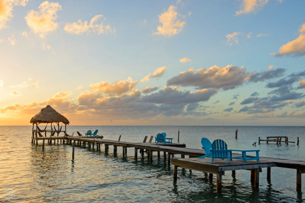 Sunrise illuminates a wooden dock with blue deck chairs and a thatched roof hut in Caye Caulker, Belize.