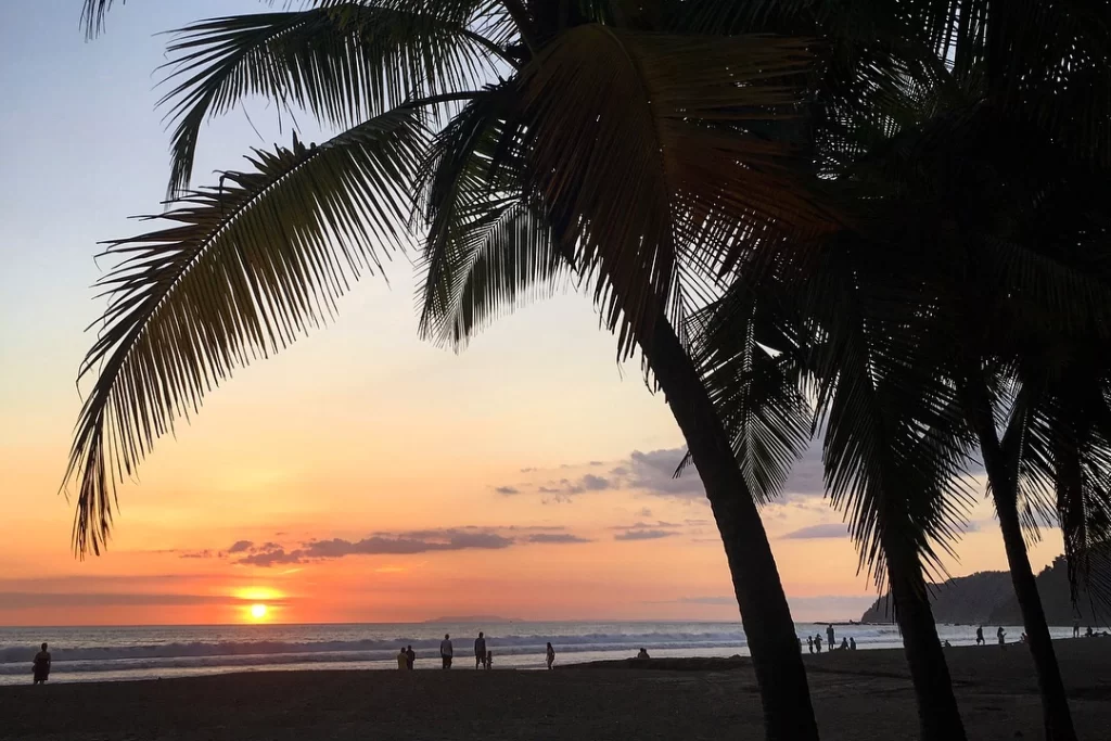 People stroll along the back-lit beach in Jaco, Costa Rica as the sun sets over the ocean.