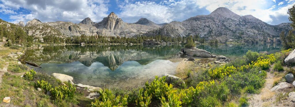 A panorama of Middle Rae Lake in Kings Canyon National Park is awash with color, with tall mountain peaks reflected in azure waters. Green corn lilies line the banks.