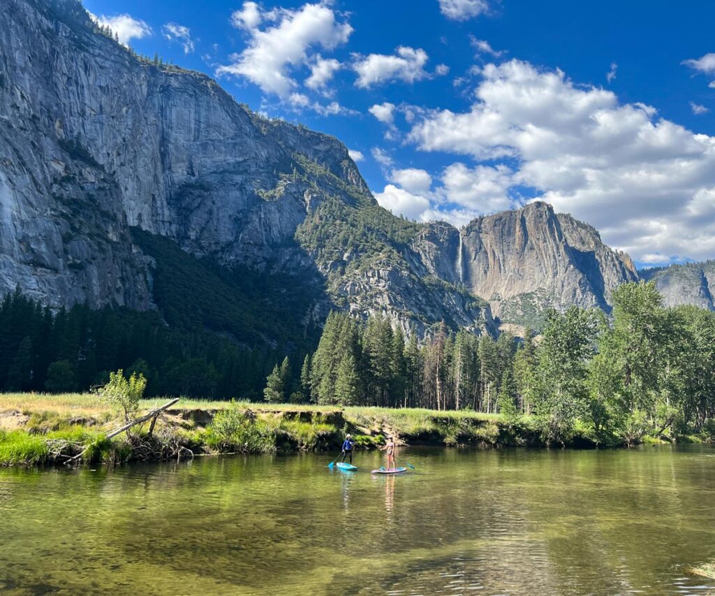 Two paddle boarders pause along the Merced River in Yosemite Valley, with Yosemite Falls in the background.