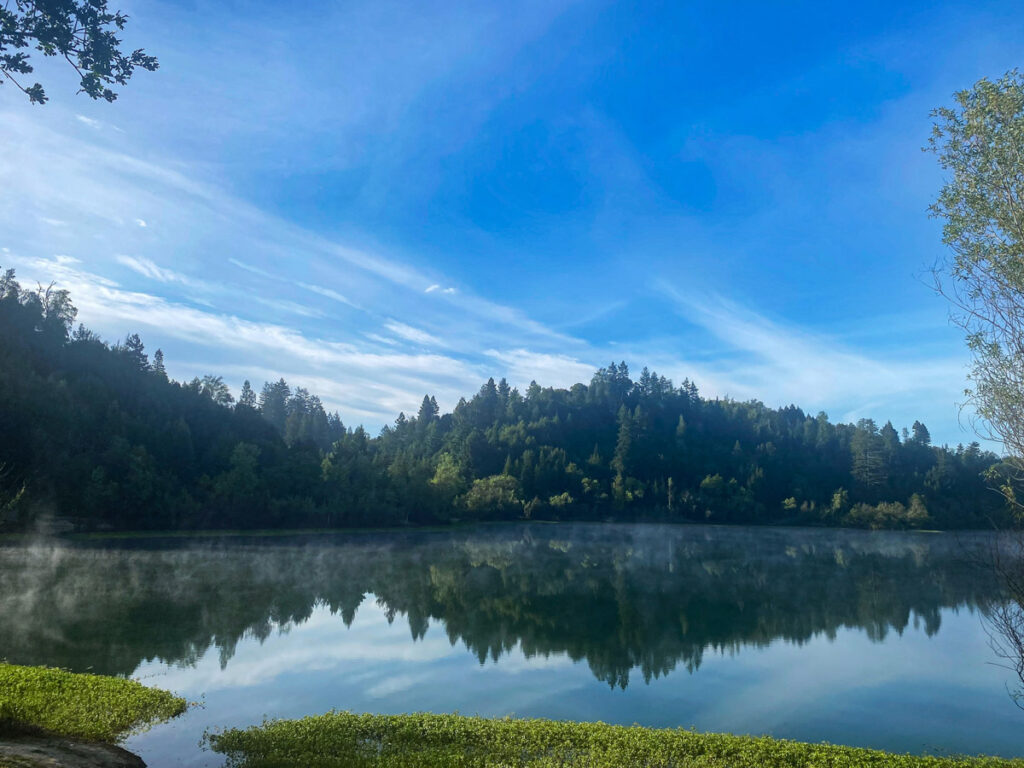 The shores of Riverfront Regional Park in Windsor, California are reflected in the waters of Lake Benoist, with lingering morning fog on a sunny day.