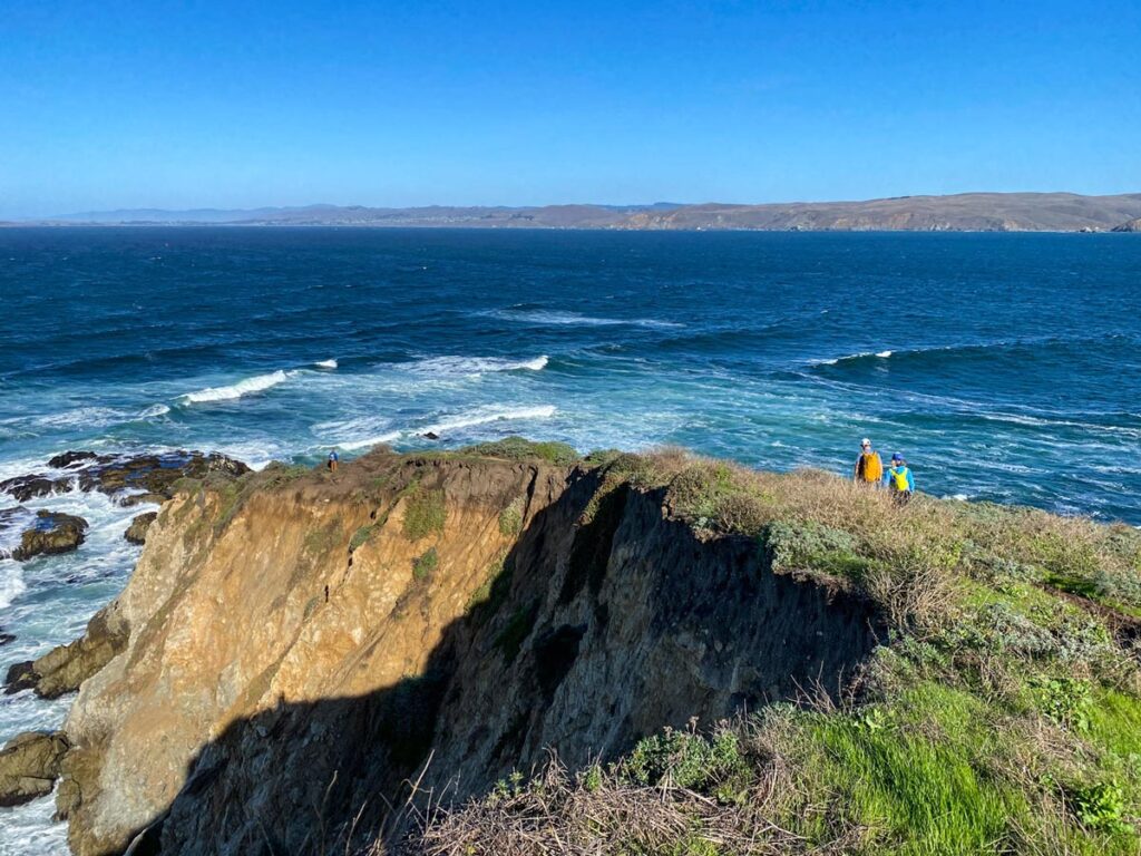 Two hikers traverse the narrow Tomales Point trail, hugging a narrow cliff with ocean on both sides, in Northern California's Point Reyes National Seashore.