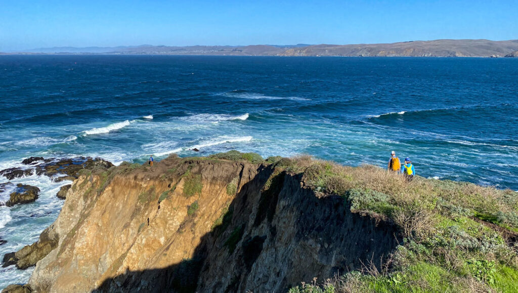 Two hikers traverse the narrow Tomales Point trail, hugging a narrow cliff with ocean on both sides, in Northern California's Point Reyes National Seashore.