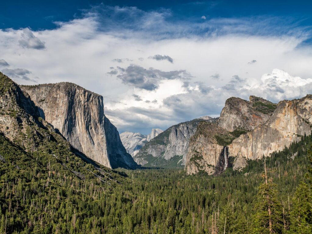 Yosemite National Park's iconic Tunnel View as seen on a partly cloudy day, with El Capitan and Half Dome prominent above the forested valley below.