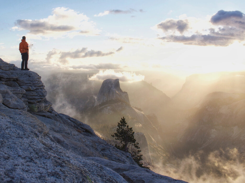 A hiker stands atop Clouds Rest in Yosemite National Park, with Half Dome shrouded by clouds in the distance.