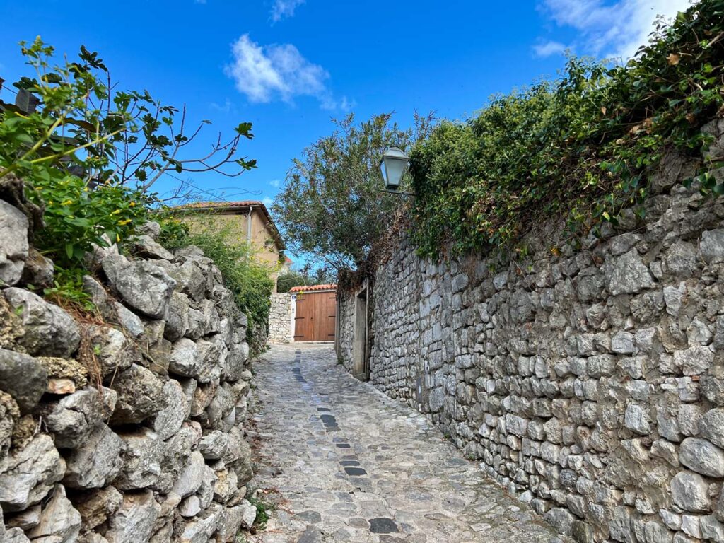 A medieval stone street winds through the old town of Krk, Croatia, with vines and olive trees lining the walls on either side.