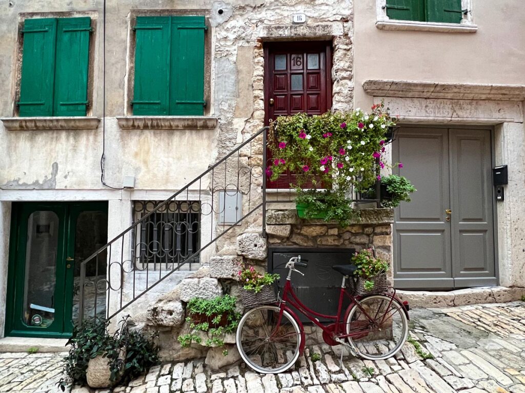 A red bicycle rests against a stone wall with potted plants in the old town of Rovinj, Croatia.