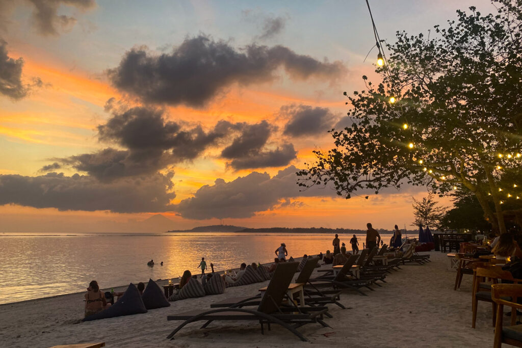 Vacationers walk the beach on Indonesia's Gili Air at sunset, with Mount Agung on Bali visible in the distance.