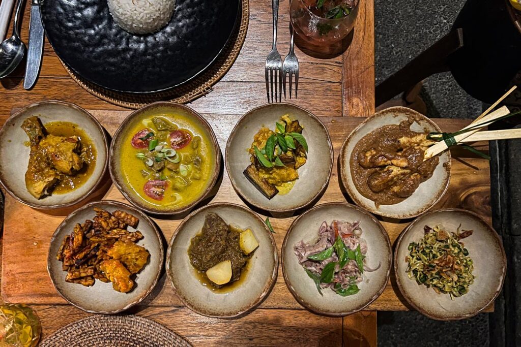 Eight dishes of traditional Indonesian cuisine are shown spread along a wooden table.