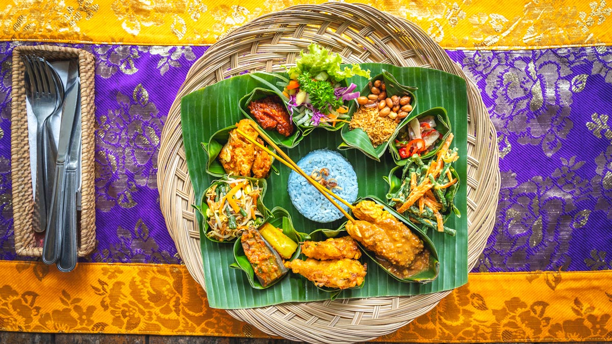 A sampler of traditional Indonesian food is laid on a banana leaf plate, on a bright yellow and purple tablecloth. Some of the best restaurants in Ubud offer creative takes on traditional fare.