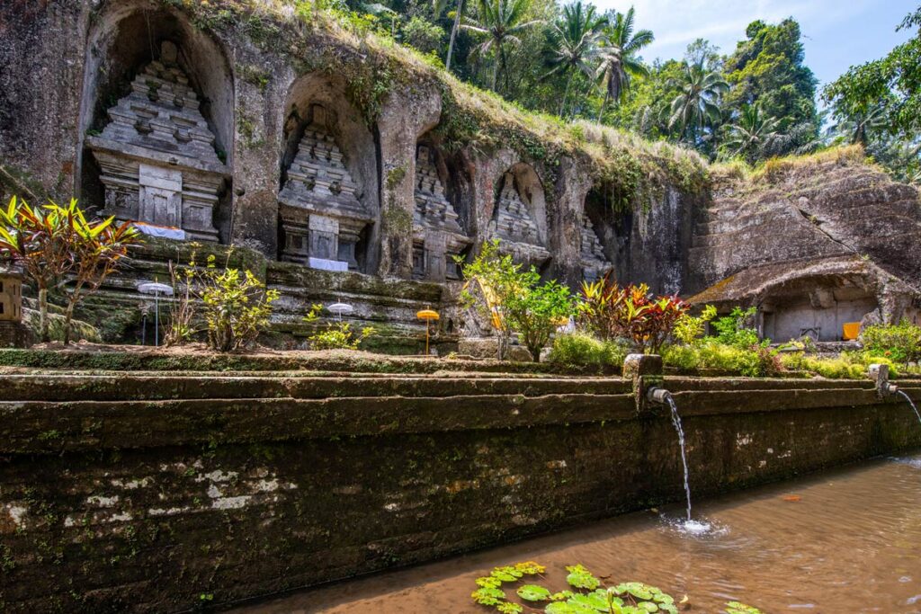 The ancient temple of Gunung Kawi near Ubud, Bali, with a series of stone temples carved into a hillside. A pond sits at the base of the temples, with palm trees and dense jungle rising on the hill above.