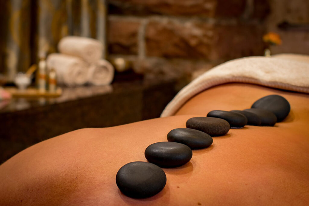 Close-up detail of black hot stones along a woman's back during a massage, with spa treatments in the background.