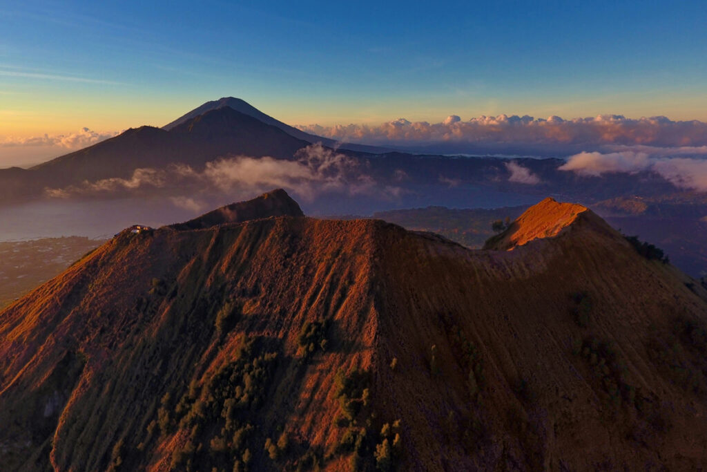 Mount Batur, on the island of Bali in Indonesia, rises above light clouds as it's bathed in the warm color tones of sunrise.