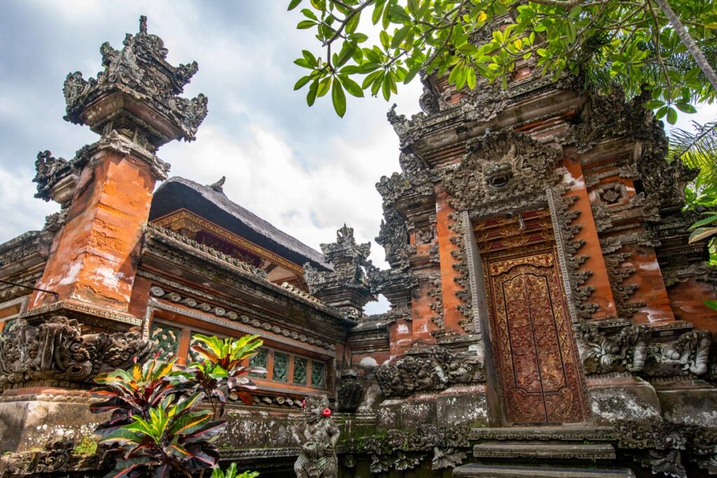 An angled fish-eye view of Saraswati Temple in Ubud, Bali, with intricate stone carvings and a Hindu motif adorning a brick entryway.