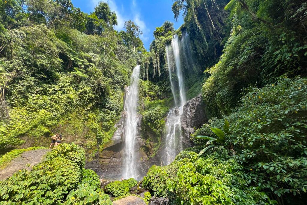 Sekumpul Waterfall is the tallest waterfall in Bali. Two figures stand on a rock at the base of the falls, appearing minute compared to the waterfall above.