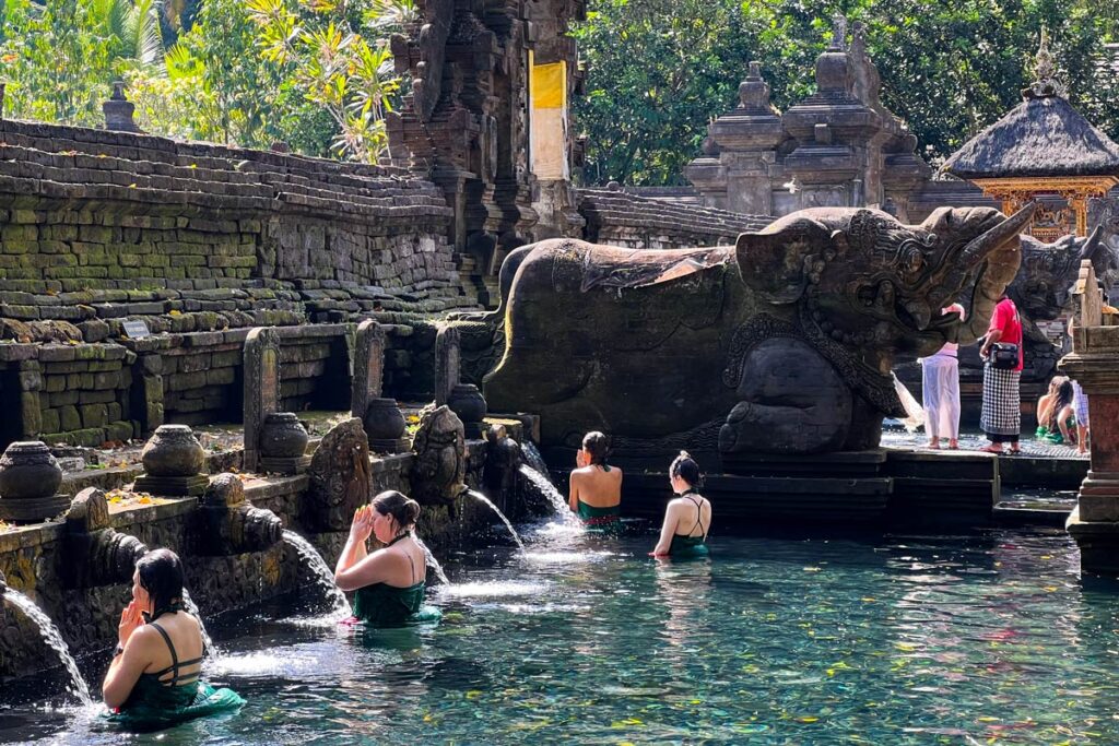 Visitors pray at the holy springs of Tirta Empul temple in Bali, Indonesia, with a large stone elephant in the background.