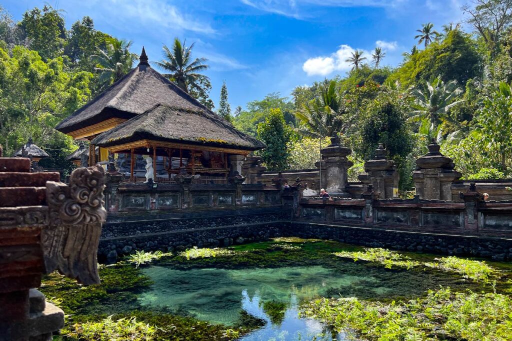 A reflecting pond at Tirta Empul in Bali, Indonesia is surrounded by a low stone wall, lush tropical vegetation, and a temple with a thatched roof.