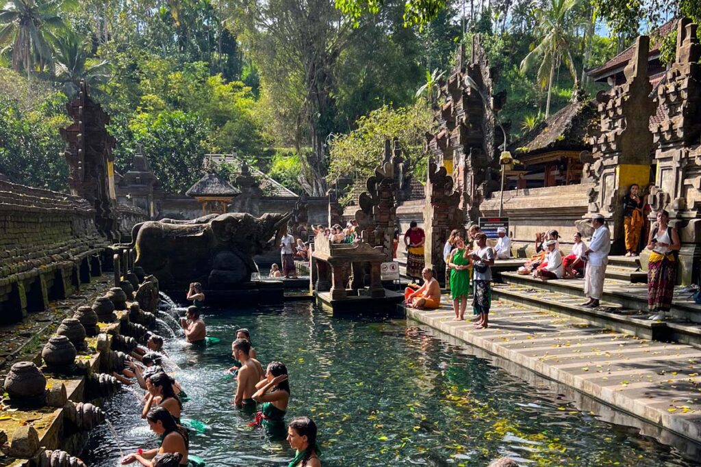 Visitors pray at the holy springs of Tirta Empul temple in Bali, Indonesia, with a large stone elephant in the background.