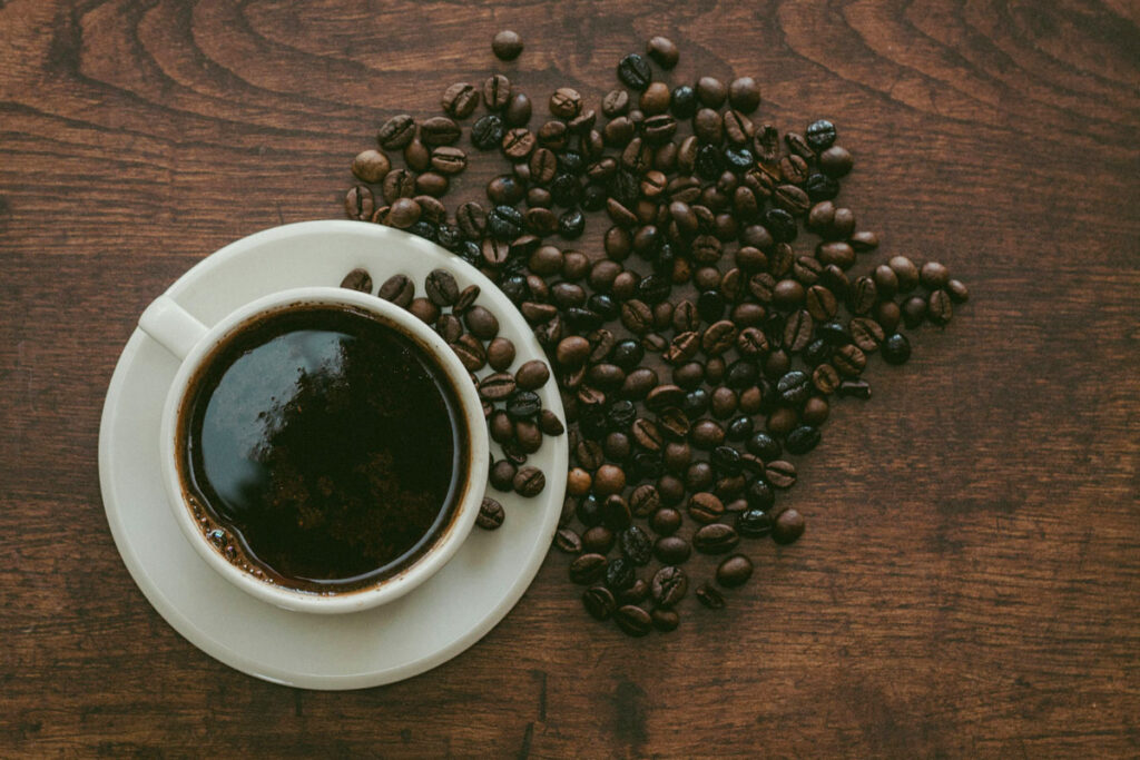 A cup of coffee is shown with roasted beans sprinkled on the dark wood table beside it. 