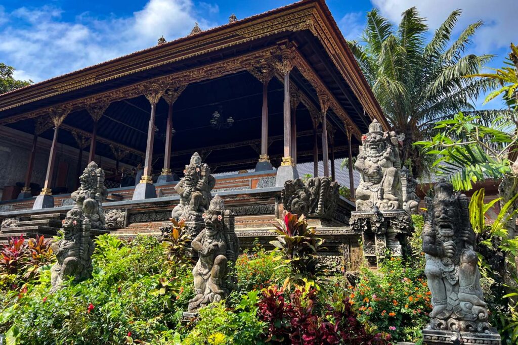 An outdoor pavilion with detailed gold carvings stands against palm fronds and Hindu stone deity statues at Ubud Palace in Bali, Indonesia.