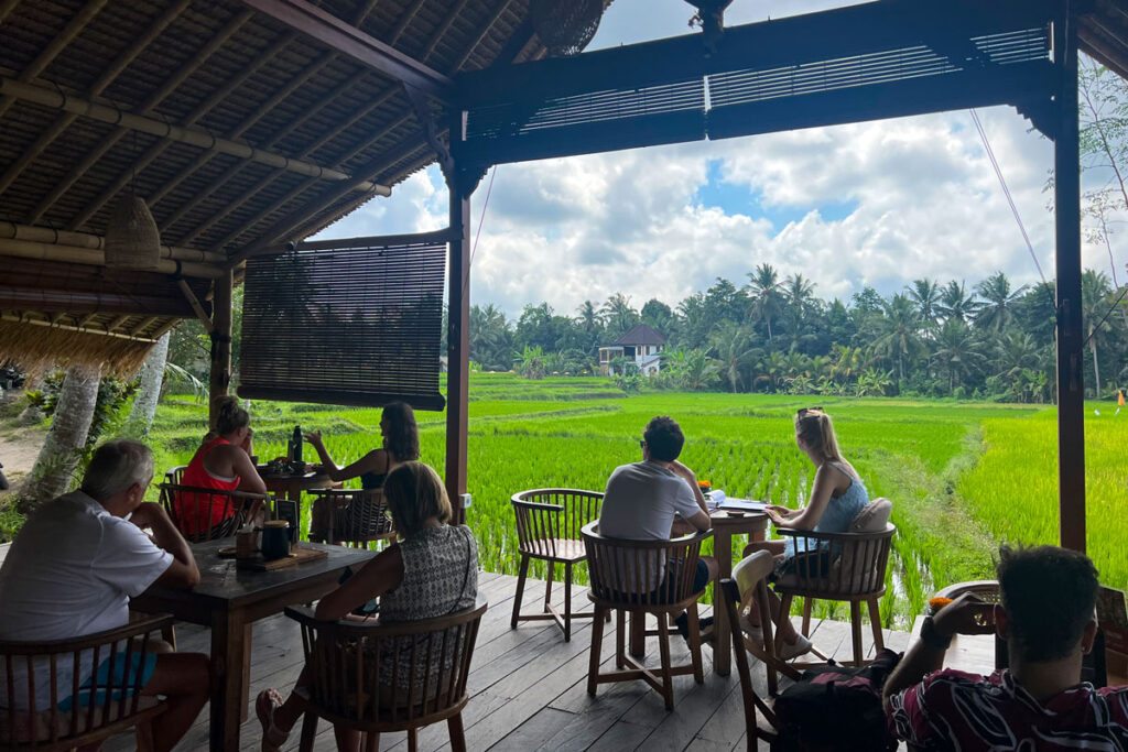 Cafe patrons sit on a covered patio, overlooking a green rice paddy and jungle in Ubud, Bali. 