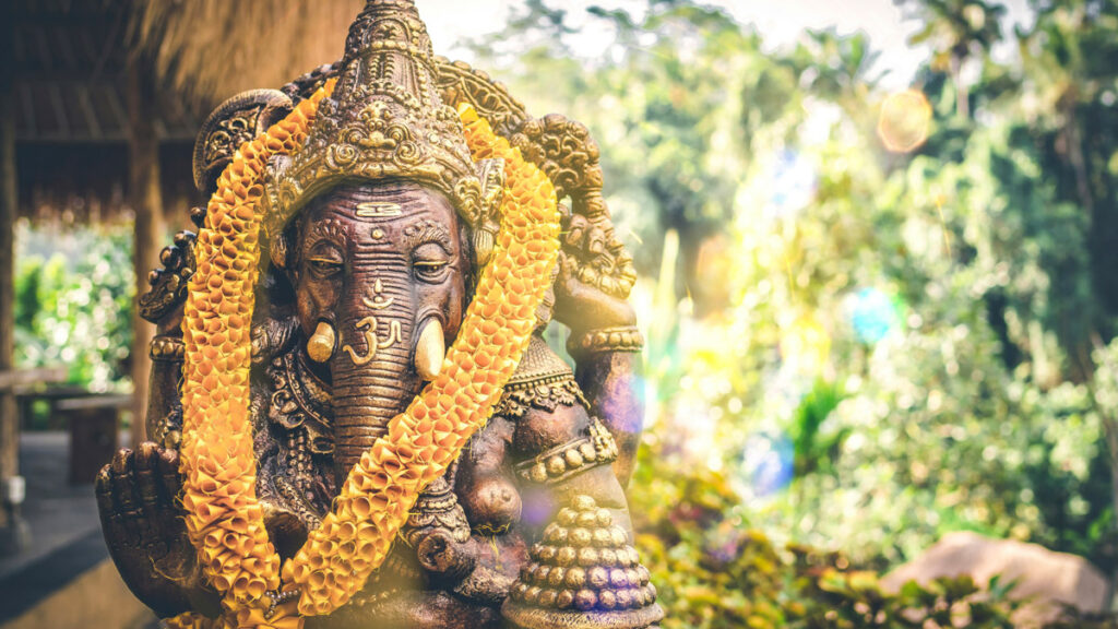 A closeup of a bronze statue of the Hindu god Ganesha, with palm fronds muted in the background.