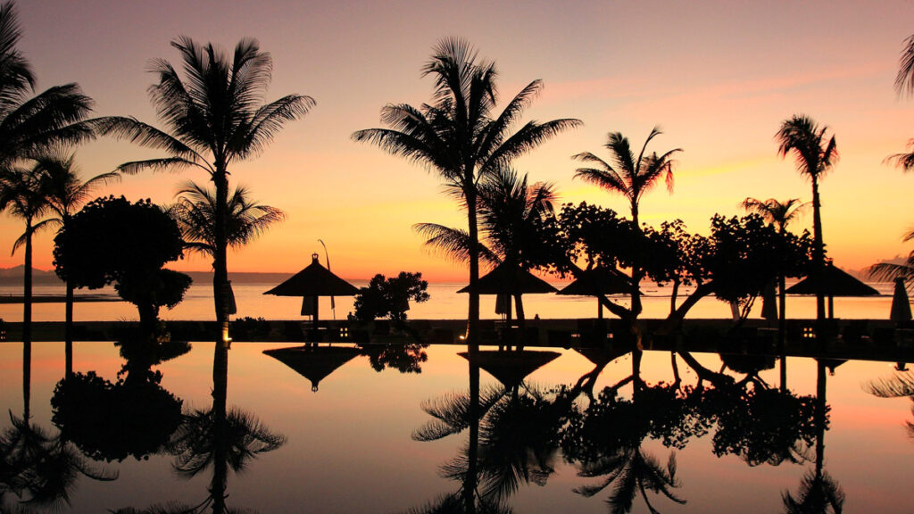 Palm fronds and small beach palapa huts are silhouetted against a sunset sky in Bali, Indonesia.