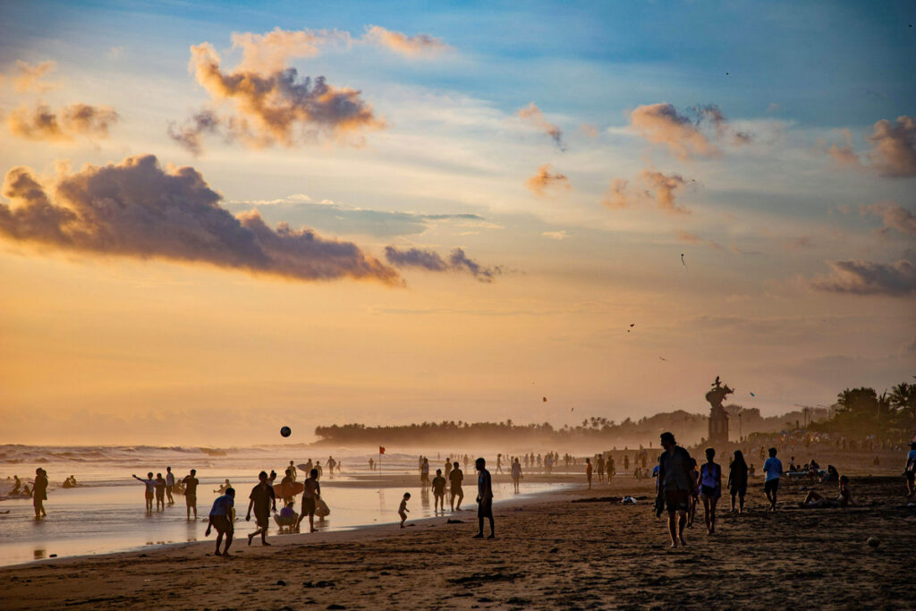 A crowded beach in Canggu, Bali at sunset, with numerous beachgoers silhouetted against the sunset sky.