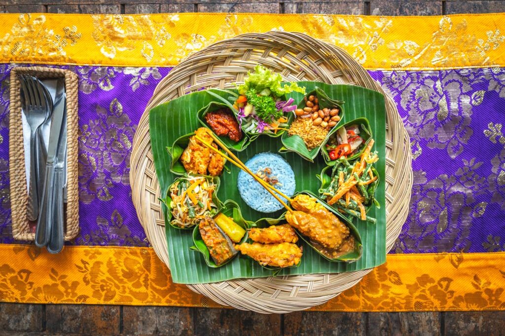 A sampler of traditional Indonesian food is laid on a banana leaf plate, on a bright yellow and purple tablecloth. 