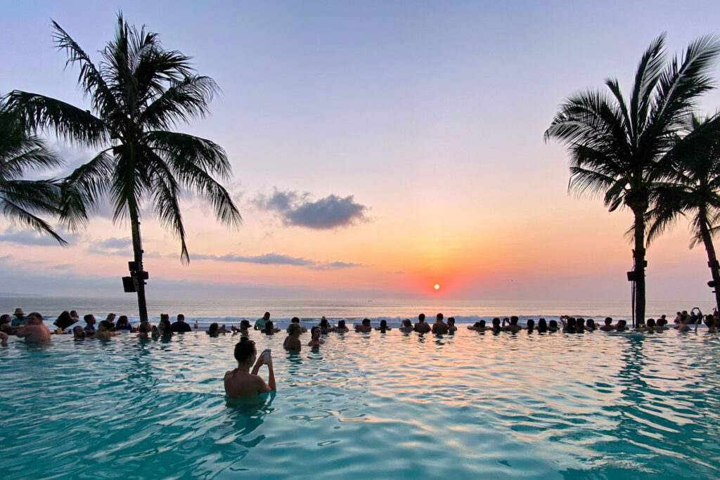 Crowds line the edge of a swimming pool, looking out to the ocean at sunset in Seminyak, Bali. Seminyak is a great place to visit on a 7 day Bali itinerary.