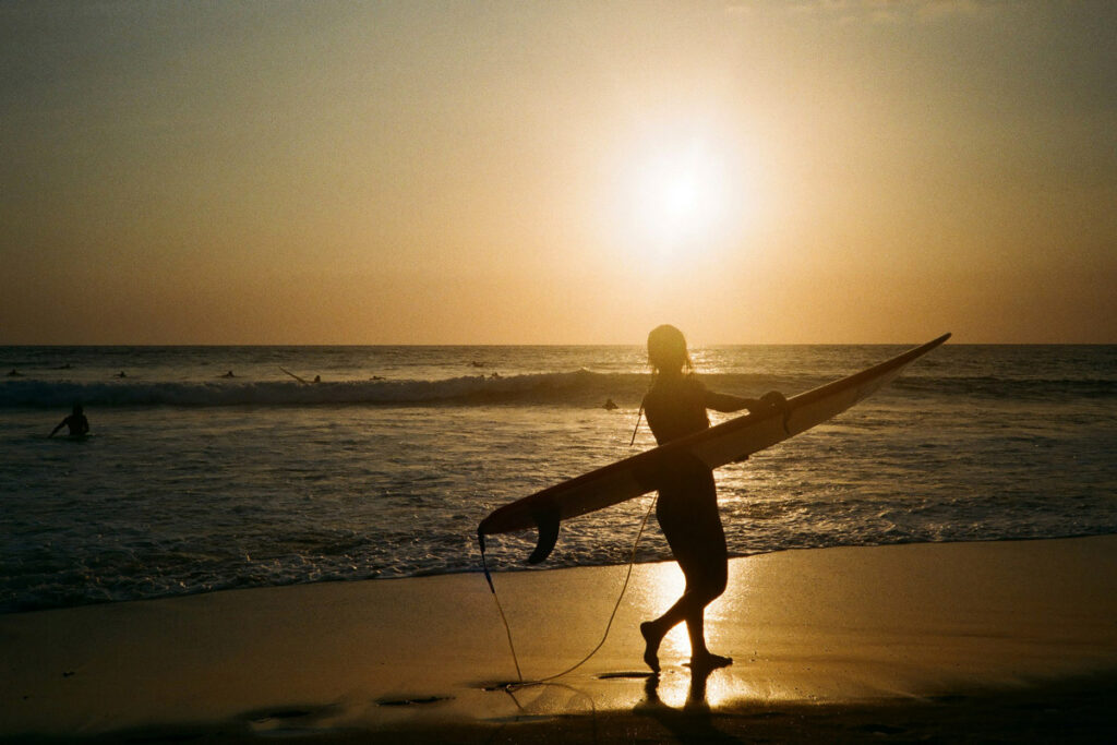 The silhouette of a woman, walking along the beach and carrying a surfboard, is accented against sunset in Canggu, Bali. When deciding where to go in Bali, Canggu is consistently a top destination for beginner surfers. 