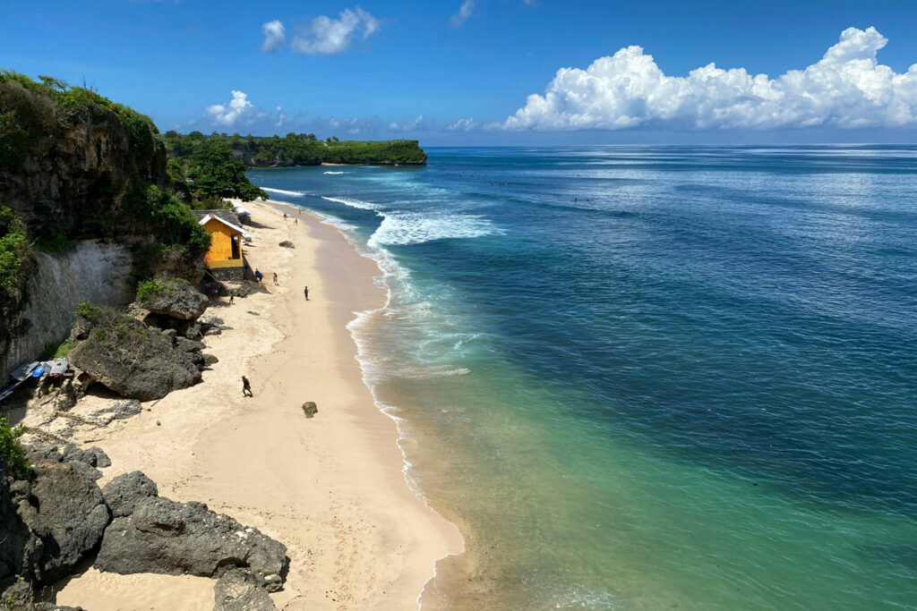 A white sand beach in Uluwatu, Bali, is seen from the cliffs above, with turquoise water and a small jungle promontory in the distance.