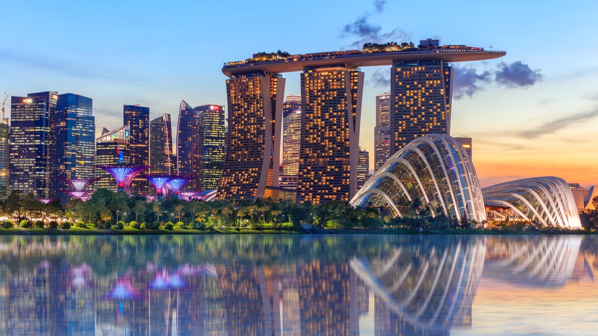 The twinkling lights of Singapore's Marina Bay Sands Hotel and Gardens by the Bay are reflected across Marina Bay at dusk.