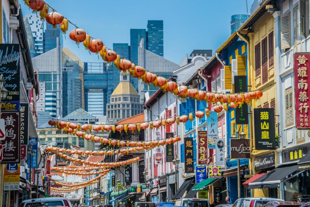 Red and yellow lanterns are strung above a street in Singapore's Chinatown neighhorbood, with traditional row buildings looking back towards modern skyscrapers