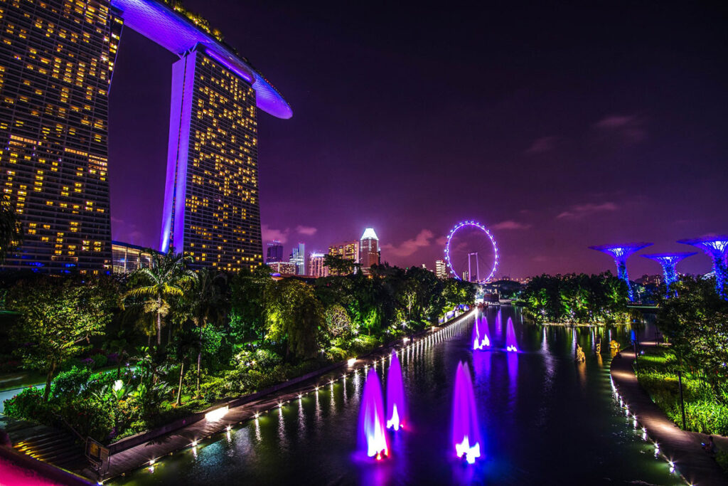 A nighttime view of Singapore's iconic Marina Bay Sands and Gardens by the Bay, both illuminated in purple lighting.