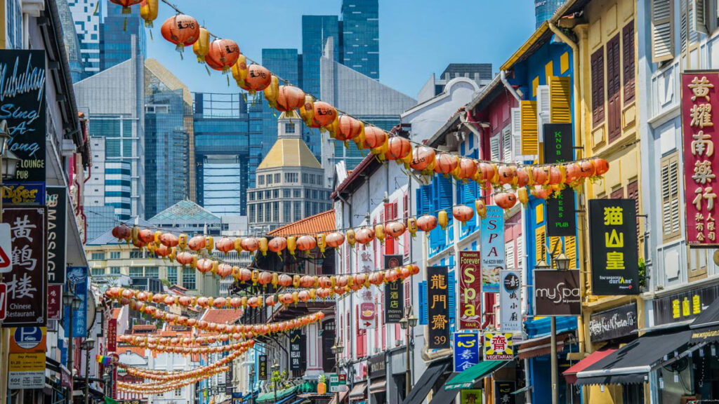 Red and yellow lanterns are strung above a street in Singapore's Chinatown neighhorbood, with traditional row buildings looking back towards modern skyscrapers