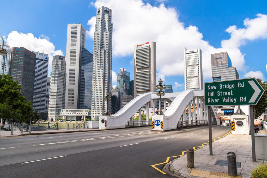 A downtown street scene in Singapore's city center shows skyscrapers and an overpass on a sunny day.