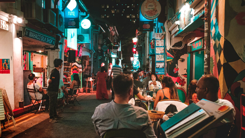 Nighttime patrons sit along a brightly-lit alleyway full of outdoor restaurants in Singapore.