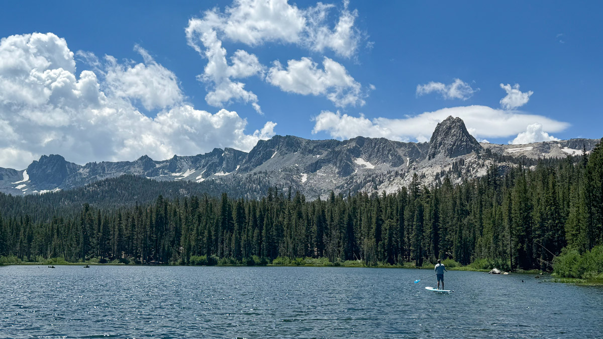 A lone paddle boarder pauses on Lake Mamie, in Mammoth Lakes, California. The lake is rimmed with tall pines and dramatic mountain peaks in the background.