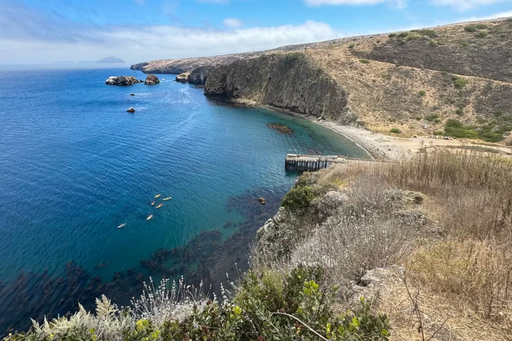 The view of Cavern Point on Santa Cruz Island in the Channel Islands, with a small group of kayakers paddling through the kelp forests far below the bluffs, and Anacapa Island visible in the distance.