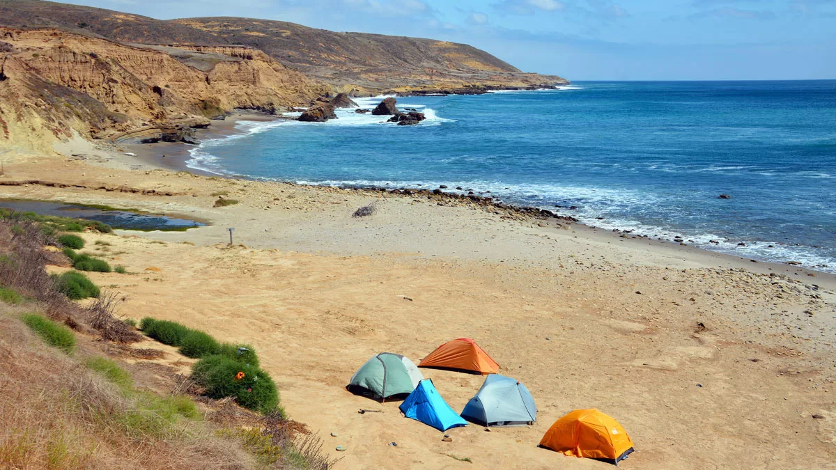 Five small tents dot the sand on a remote coastline of Santa Rosa island, in Channel Islands National Park. Waves break along the rocky cliffs in the background.