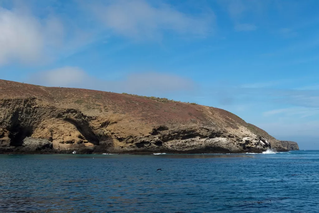 The coast of Santa Barbara island, part of Channel Islands National Park, is seen from offshore, with small waves breaking along the dry cliffsides.