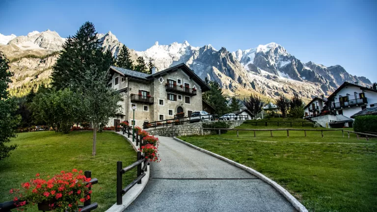 A traditional Alpine building, with red geraniums in window flower boxes, sits against a backdrop of snowcapped peaks in Courmayeur, Italy.