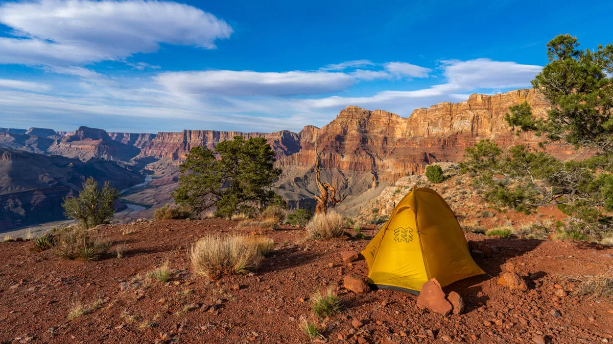 A solitary yellow tent sits on a red dirt outcropping overlooking the Grand Canyon. The Colorado River is visible in the distance, snaking among the red plateaus.