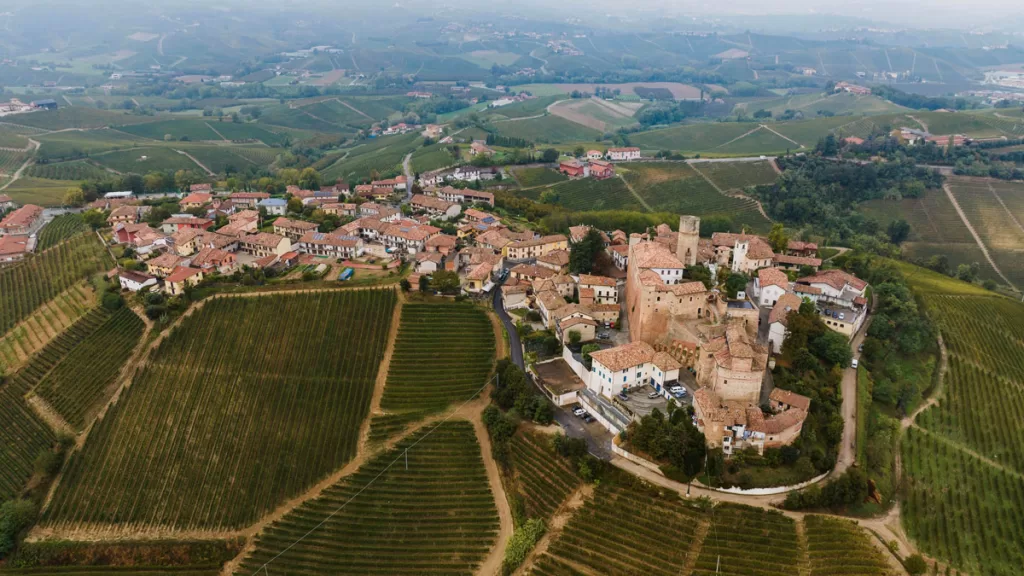 The medieval stone town of Castiglione Falletto in Piedmont, Italy, stretches along a green hillside, surrounded by vineyards.