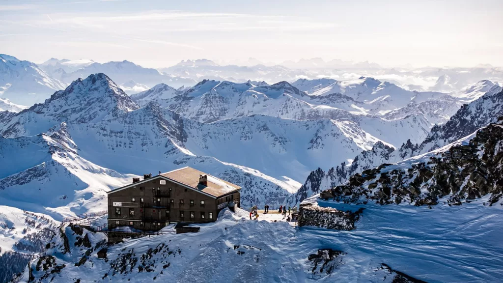 A traditional Alpine lodge sits on a mountainside near Courmayeur, Italy, surrounded by snowy mountain peaks.