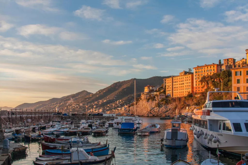 Small boats dot the harbor in Camogli, Italy, as the setting sun illuminates the buildings on the cliffside beyond.