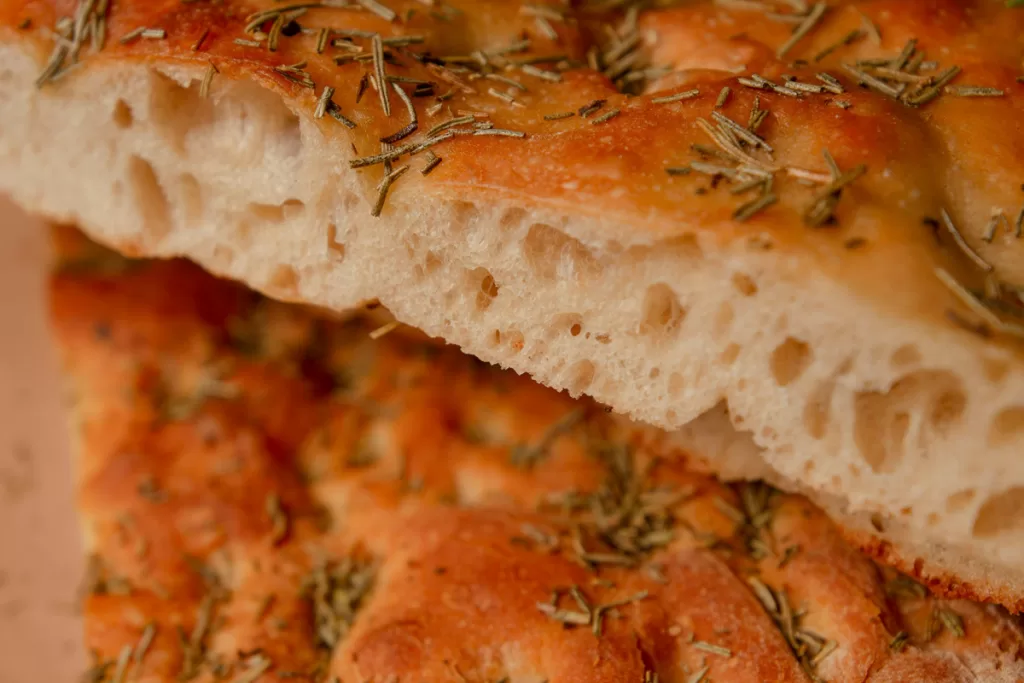 A textured close-up of focaccia bread, with dried rosemary sprinkled on the crisp crust.